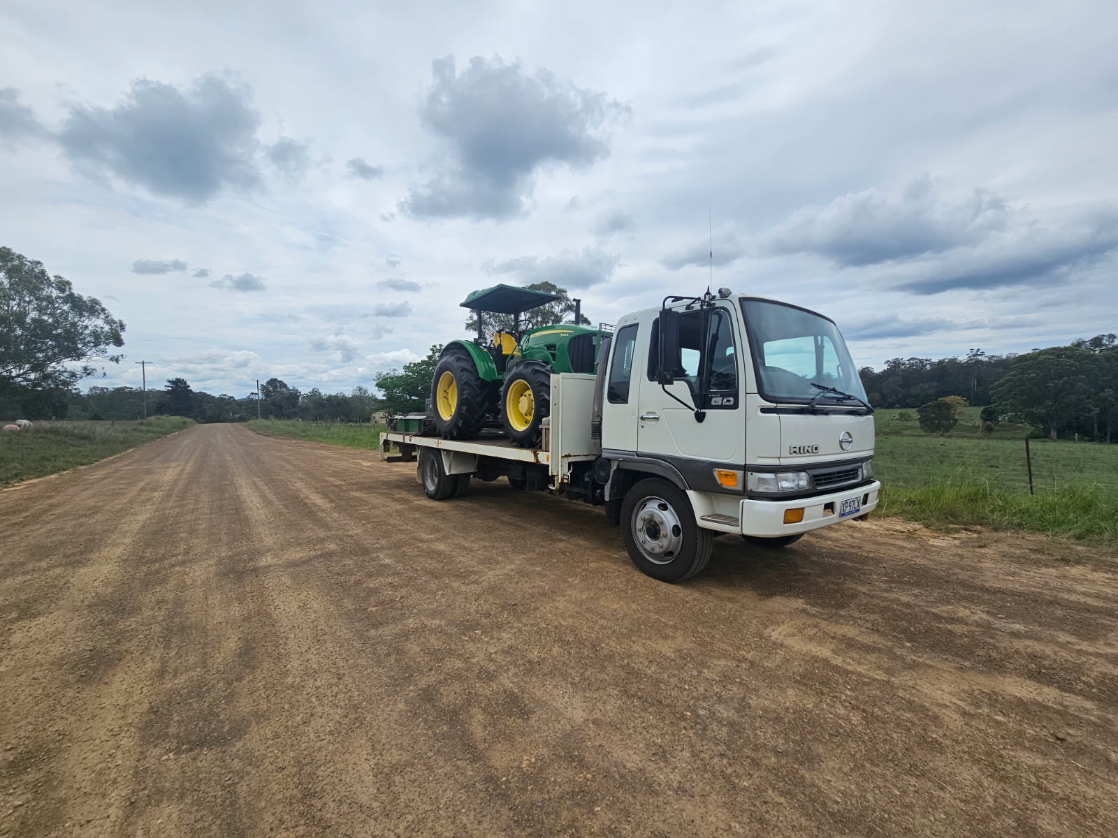 A white flatbed lorry carrying a green farm tractor is parked on a wide, unpaved rural road surrounded by trees and fields under a cloudy sky.
