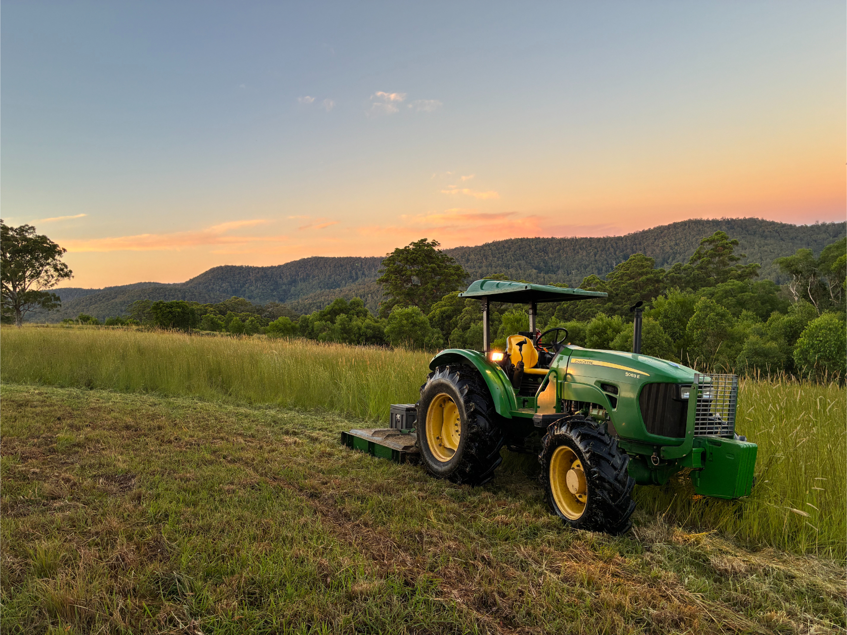 A green tractor sits in a grassy field at sunset, surrounded by tall grass and rolling hills with trees. The sky is colourful, with shades of orange, pink, and blue above the distant mountains.