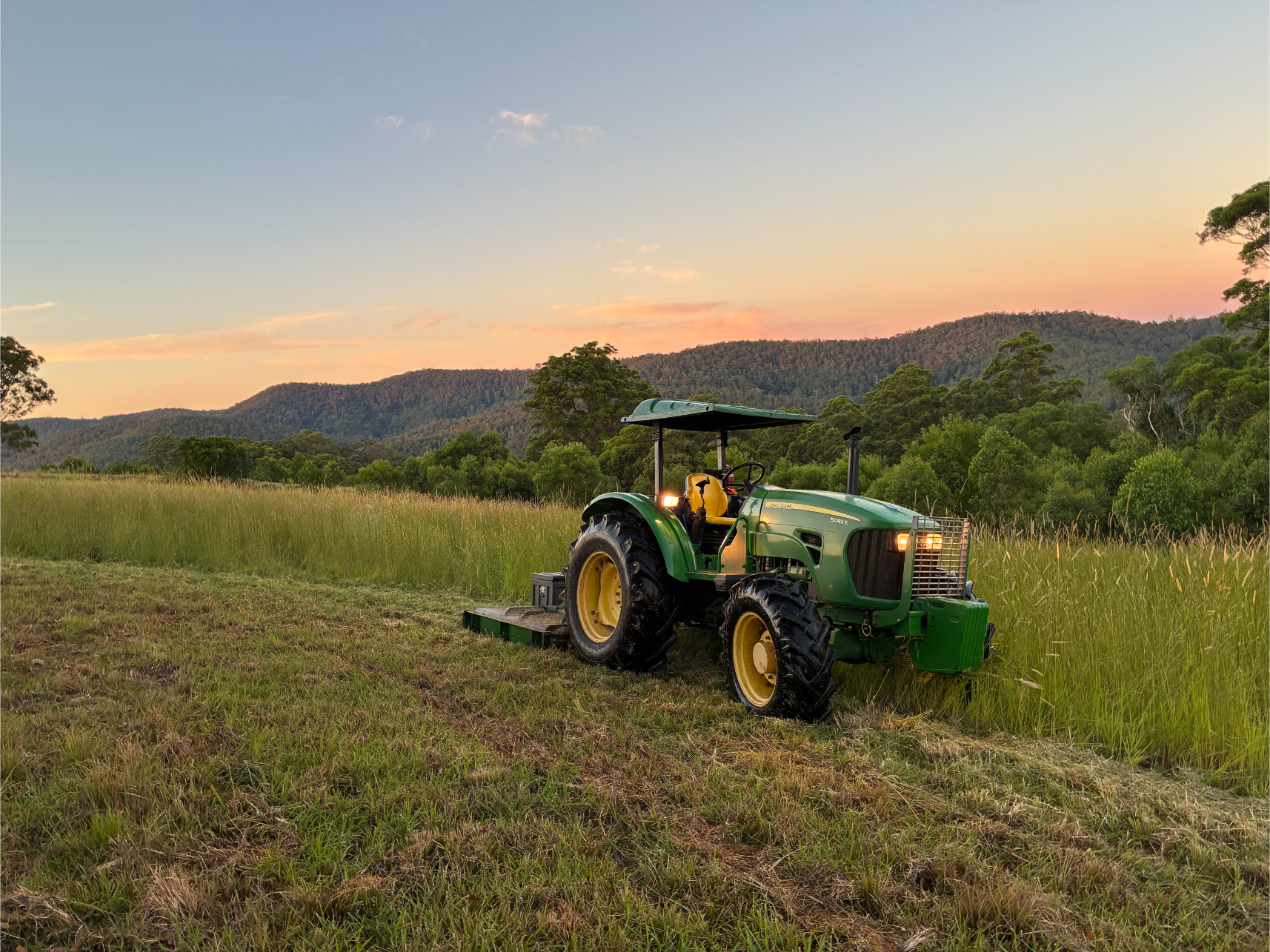 A green tractor sits on a grassy field at sunset, surrounded by tall grass and trees, with hills and a colourful sky in the background.