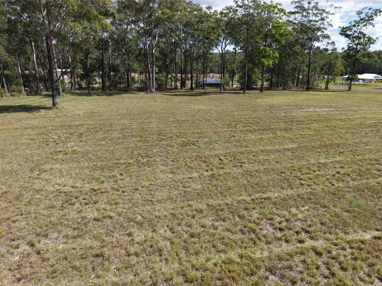 A grassy, open field with patchy grass, surrounded by tall trees—ideal for paddock slashing Mid Coast NSW. In the background, a few houses are partially visible through the trees under a partly cloudy sky.