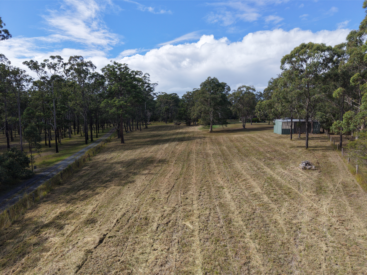 Service area featuring a large mowed field, surrounding trees, shed, and blue sky with white clouds. Aerial view.