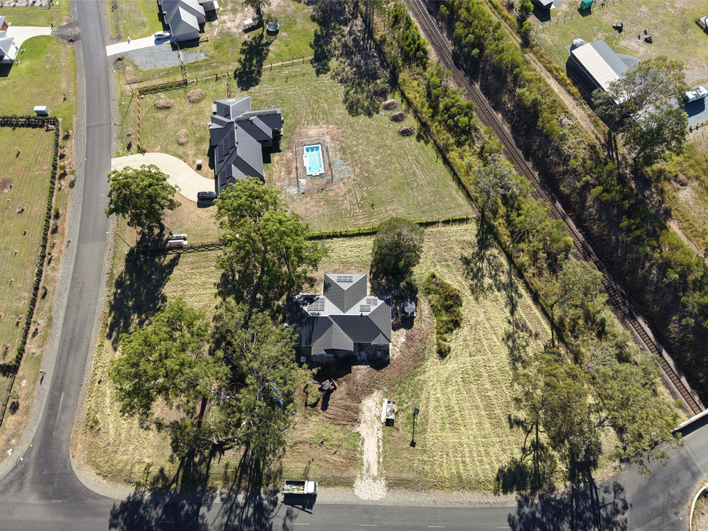 Aerial view of a suburban property with a house, front and back gardens, a swimming pool, trees, and surrounding roads. Nearby houses, railway tracks, and open grassy areas are also visible.